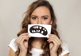 A young woman holding up a drawn-on smile with missing and misshapen teeth